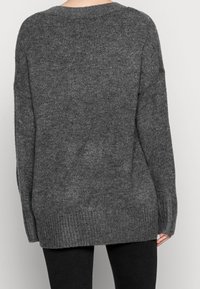 Person, der einen lockeren, dunkelgrauen Strickpullover mit langen Ärmeln trägt, von hinten vor einem schlichten hellen Hintergrund gezeigt.