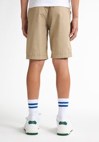 Pantalones cortos de algodón beige con dobladillos enrollados, combinados con calcetas blancas con rayas azules y zapatillas blancas con acentos verdes.