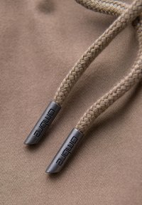 Cordons de serrage tressés beiges avec embouts métalliques gravés "Ombre" sur fond de tissu beige texturé.