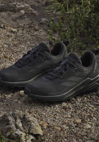 Een paar zwarte Adidas Terrex trailrunning schoenen op rotsachtige grond met kleine groene planten op de achtergrond.