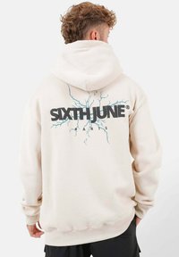 Beige Kapuzenpullover mit großem schwarzem Logo "SIXTHJUNE" und blauem Blitzdesign auf dem Rücken. Verfügt über eine geräumige Kapuze und gerippte Bündchen.