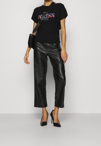 Schwarzes T-Shirt mit kurzen Ärmeln und farbenfrohem Logo, kombiniert mit hoch taillierter, glänzender schwarzer Lederhose und schwarzen spitzen Pumps.