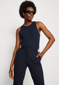 Navy-blaues, geripptes Tanktop mit figurbetontem Design, kombiniert mit hoch taillierten, navy-blauen Hosen mit weißen Streifenakzenten an der Taille. Trägt schwarze Sonnenbrillen.