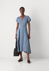 Lichtblauwe midi-jurk met een V-hals, korte pofmouwen, een geraffineerde taille en een geplooide rok. Gecombineerd met zwarte blokkengeneratie muilen en een zwarte tas.