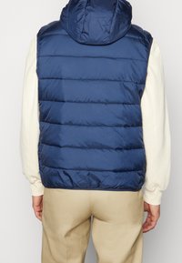 Tmavě modrá prošívaná vesta s kapucí, s horizontálními prošívanými sekcemi, hladkou texturou a elastickými otvory na paže, nošená přes světlé tričko.