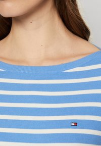 Zbliżenie na szyję i ramię kobiety ubraną w niebiesko-biały sweter w stylu boatneck w paski, z małym logo w kolorach czerwonym, białym i czarnym.