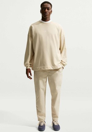 Mężczyzna w beżowej bluzie i spodniach Nike oraz ciemnych butach sportowych, stojący na jednolitym białym tle, ręce swobodnie opuszczone wzdłuż ciała.
