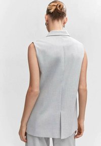 Žena s blond vlasy v drdolu, nosící stříbrné náušnice ve tvaru kruhu a bezrukávový světle šedý blejzer, zobrazená zezadu na jednobarevném pozadí.