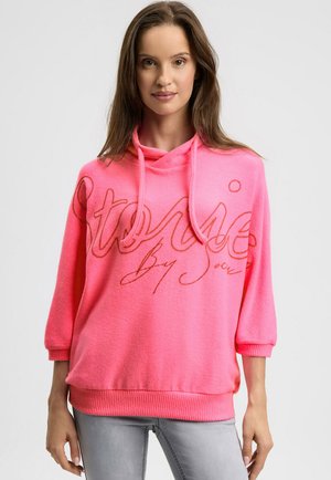 Frau trägt einen leuchtend pinken Kapuzenpullover mit orangefarbener, kursiver Schrift, kombiniert mit hellgrauen, eng anliegenden Jeans, steht vor einem einfachen Hintergrund.