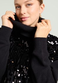 Suéter de punto negro con un cuello alto de canalé. Presenta un frente adornado con lentejuelas y puños de canalé para añadir textura y detalle.