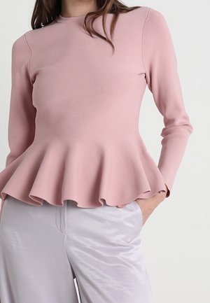 Femme portant un haut péplum rose clair à manches longues associé à un pantalon en satin gris clair, debout avec une main dans la poche.