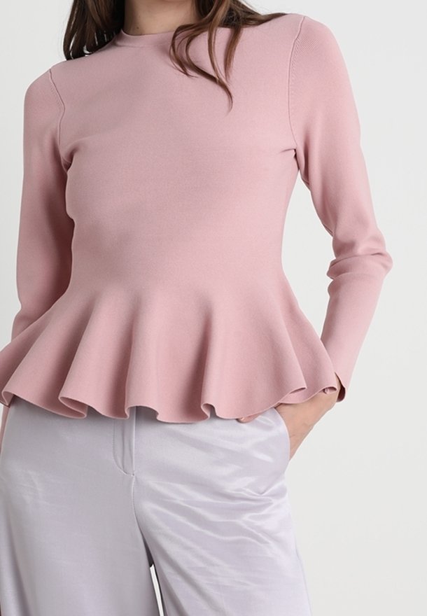 Femme portant un haut péplum rose clair à manches longues associé à un pantalon en satin gris clair, debout avec une main dans la poche.