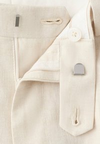 Close-up of beige linen trousers showing button, metal hook and bar fasteners, zipper fly, and stitched buttonholes.