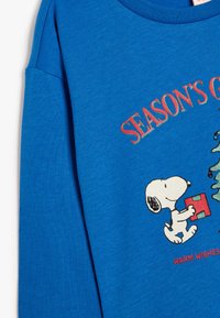 Blaues Langarmhemd aus Baumwolle. Mit einem Grafikdesign von Snoopy, der ein Geschenk trägt, sowie dem Text "Saisonale Grüße" und "Herzliche Wünsche".