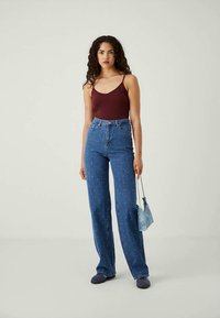 Kvinde med krøllet hår iført en bordeaux farvet tanktop, højtaljede blå jeans med hvide prikker, mørke sko og holder en lille blå håndtaske.