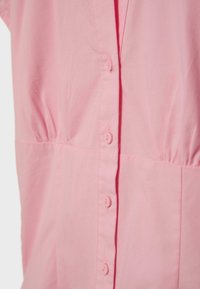 Nærbillede af en lyserød skjorte med lodrette knapper og rynket stofdetalje nær taljen på en ensfarvet baggrund.