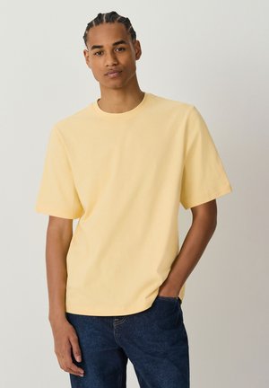 Young man with braided hair wearing a pale yellow T-shirt and dark blue jeans standing against a plain light background.