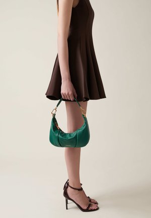 Borsa in pelle verde acqua con accessori dorati, forma semi-circolare, tenuta da una persona in un vestito marrone, in piedi su sandali con tacco alto.