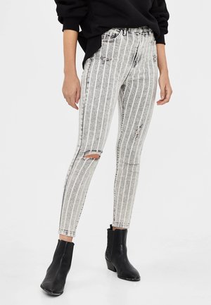 Jeans skinny taille haute en denim gris avec des rayures verticales blanches, présentant des détails déchirés et une coupe près du corps. Associés à des bottines noires.