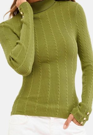 Pull vert côtelé en maille avec motifs torsadés, col montant et boutons dorés aux poignets, porté avec un pantalon blanc.