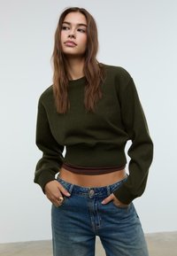 Giovane donna con lunghi capelli castani, che indossa un maglione corto verde scuro sopra una camicia marrone e jeans blu, con le mani in tasca, che guarda di lato.