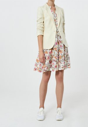 Kvinde iført en cremet blazer over en blomsterkjole til knæene med hvide sneakers, stående mod en ensfarvet hvid baggrund.