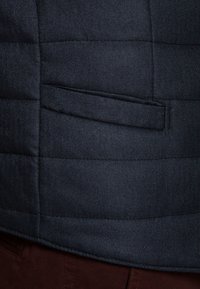 Granatowa pikowana kamizelka o strukturze w jodełkę, z pojedynczą kieszenią po lewej stronie, zestawiona z brązowym materiałem poniżej.