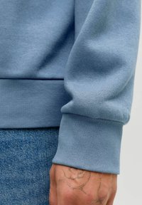 Ανοιχτό μπλε φούτερ με ριμπ σαγκάνια και τελείωμα, κατασκευασμένο από ένα απαλό βαμβακερό μείγμα, με λεία υφή και ελαφριά γυαλάδα.