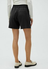 Zwarte shorts van zacht materiaal, met een brede elastische tailleband en twee achterzakken, gestyled met beige loafers.
