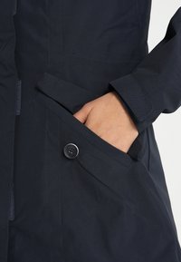 Marinblå vattentät jacka med knappstängning, sidofickor och justerbara ärmar; slät textur och minimalistisk design.