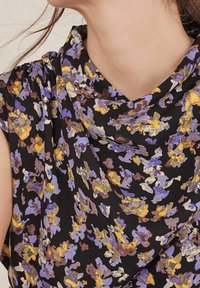 Czarna bluzka w kwiatowy wzór z purpurowymi, żółtymi i brązowymi abstrakcyjnymi motywami, wykonana z lekkiego materiału, z dekoltem w kształcie drapowania. Widziane krótkie rękawy.