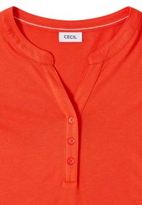 Chemise Henley orange vif avec trois boutons et un col rond présentant une petite fente, étiquetée "CECIL" à l'intérieur du col.