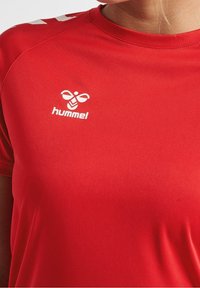 Punainen urheilullinen T-paita, joka on valmistettu sileästä kankaasta. Siinä on pyöreä pääntie, lyhyet hihat ja valkoinen logo rintaosassa.