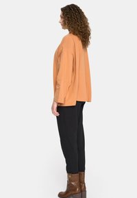 Luźna, pomarańczowa bluzka z długim rękawem zestawiona z czarnymi spodniami i brązowymi botkami za kostkę. Gładka faktura i prosty design. Widok z boku.