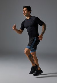 Svart kortermet treningstopp kombinert med svarte shorts med blå detaljer. Modellen løper iført svarte treningssko.