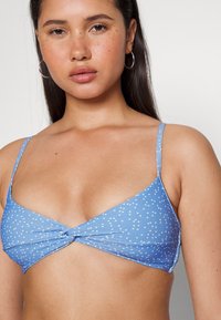 Top de bikini azul com pequenas bolinhas brancas. Apresenta um design de frente torcido e alças finas ajustáveis. Textura de tecido suave.