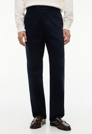 Mand iført marineblå bukser, hvid sweater med manchetlogo og mørkebrun læderloafers stående mod hvid baggrund.