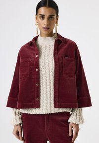 Bordeaux corduroy jack met twee voorzakken en zilveren drukknoopsluitingen, gelaagd over een crèmekleurige ribgebreide trui.