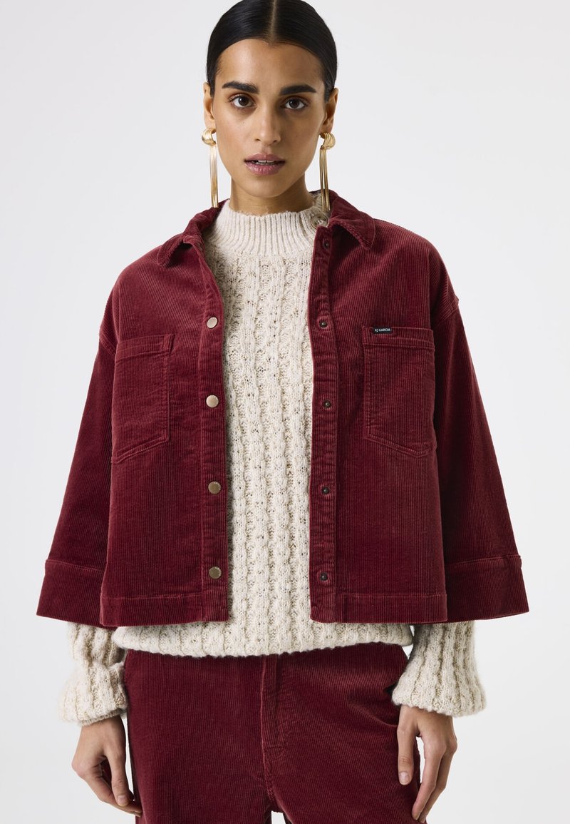 Bordeaux corduroy jack met twee voorzakken en zilveren drukknoopsluitingen, gelaagd over een crèmekleurige ribgebreide trui.