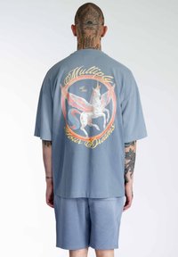 Blå oversized t-shirt med et farverigt enhjørning grafik og teksten "Multiplicer Dine Drømme" samt et etableringsår, iført med matchende shorts.