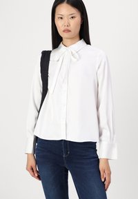Blouse blanche à manches longues avec un col en nœud et une fermeture à boutons. Tissu lisse, design légèrement court, associé à un jean bleu foncé.
