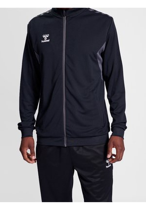 Mann trägt eine schwarze Sportjacke mit Reißverschluss und passende schwarze Hose mit weißem hummel-Logo auf Brust und Oberschenkel.
