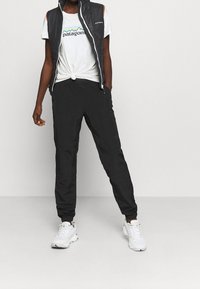 Svart vadderad väst över en vit t-shirt med grafisk design av berg och svarta byxor. Vita sneakers fulländar utomhusoutfiten.