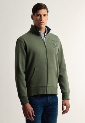 Sudadera de cremallera color verde oliva en suave material con un cuello alto, que presenta un logo en el pecho y bolsillos frontales; puños y dobladillo de canalé.