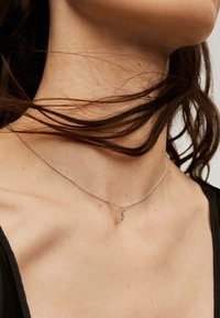 Collarín de cadena de plata reposando sobre la clavícula, que presenta un pequeño colgante en forma de letra "P".