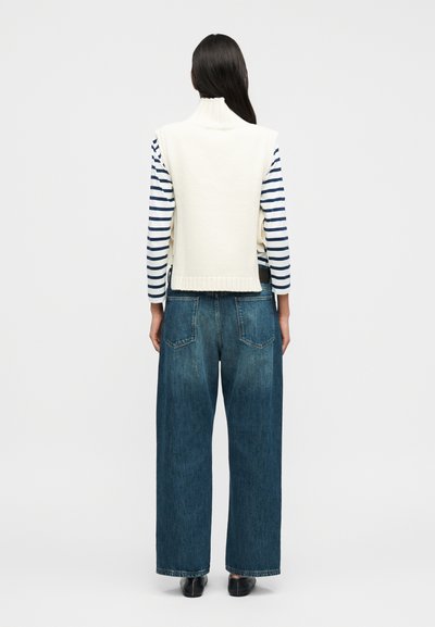 Λευκό πουλόβερ χωρίς μανίκια πάνω από μπλε-σκούρο και άσπρη ριγέ μακρυμάνικη μπλούζα, σε συνδυασμό με τζιν παντελόνι με φαρδιά μπατζάκια. Απλός σχεδιασμός; χαλαρή εφαρμογή.