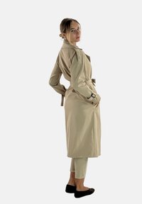 Beige trenchcoat med bältesmidja, sidfickor och långa ärmar. Slätt tyg med lätt figursydd passform och detalj med bakre slits.