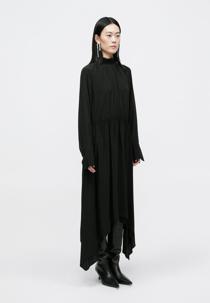 Un vestido negro de manga larga con cintura fruncida, dobladillo asimétrico y mangas anchas. Combinado con botas negras de punta. Textura suave.