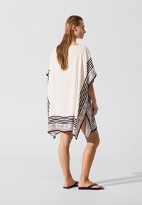 Kremasti kaftan s crnim prugama na rubovima i jedinstvenim uzorkom. Kratki rukavi, širok kroj i bočni prorezi omogućuju lako kretanje.