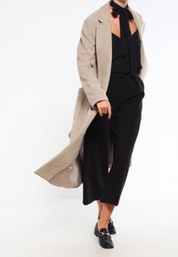 Beige long coat with a tailored design, black top, and wide-leg black trousers. Black loafers with gold hardware accents.